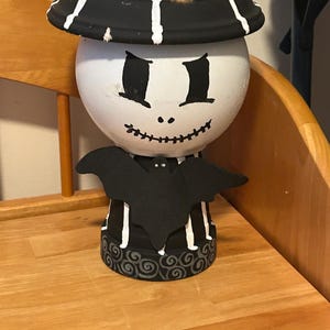 Peut inclure: Pot décoratif peint à la main sur le thème d'Halloween. Le pot est blanc avec un chapeau noir et un nœud papillon en forme de chauve-souris. Le visage a des yeux noirs et un sourire cousu. La base a un motif tourbillonnant noir et blanc.