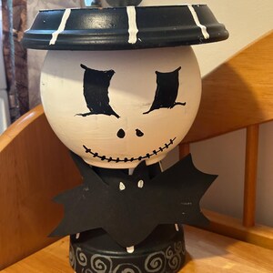 Peut inclure: Objet décoratif sur le thème d'Halloween avec un visage peint, des yeux noirs et un sourire cousu. Il possède un chapeau noir à rayures blanches et un motif en forme de chauve-souris. La base est noire avec des volutes blanches.