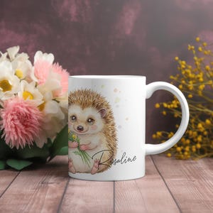 Pode incluir: Caneca de cerâmica branca com uma ilustração em aquarela de um ouriço-cacheiro fofo segurando flores. A caneca tem o nome "Rosaline" em fonte cursiva. O fundo inclui flores e uma superfície de madeira.