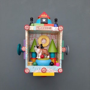 May include: A colorful wooden box with a map background and the word "PLAYTIME" on the front. Inside the box, there are two toy dogs, a red ball, and a green tree. The box is decorated with numbers and other colorful details.