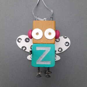 May include: A wooden robot ornament with wings, painted in brown, teal, white, and pink. The robot has large silver eyes and a letter "Z" on its chest.