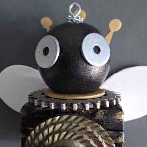 May include: A decorative figurine featuring a dark brown spherical head with two silver washer eyes and gold antennae. The body is a dark block topped with a metallic gear, featuring a large gold circular accent and white wings extending from the sides.