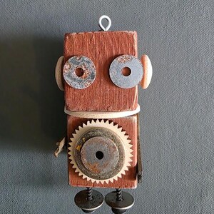 May include: A handcrafted wooden robot sculpture with a reddish-brown body. The robot features metal washers for eyes, a gear for a torso, and metal hardware for feet. A wire loop is attached to the top for hanging. The robot is a unique piece of art.