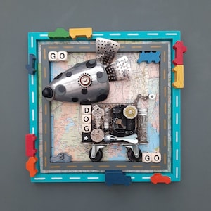 May include: A framed mixed media artwork featuring a whimsical robot dog made of metal, gears, and other found objects. The dog is on a road with the words "GO" and "DOG" on the sides. The frame is painted turquoise with a yellow, green, and red border.