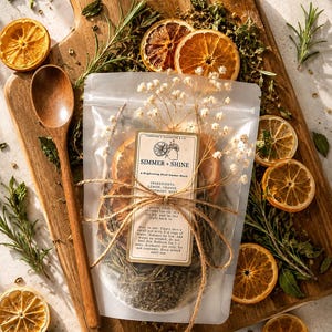 May include: A translucent pouch labeled Simmer + Shine containing dried orange slices, rosemary, and mint, tied with twine and topped with dried baby's breath. A wooden spoon rests beside the pouch, which features a list of ingredients.