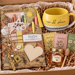 May include: A gift box containing a wrapped book with a reading list, a yellow mug with the text rise & shine, three tea packets labeled Black Tea, Earl Grey, and Herbal Tea, a small jar of honey with a wooden dipper, and a metal sunflower bookmark.