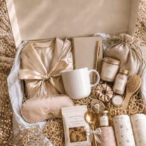 May include: A gift box containing a champagne-colored satin robe and eye mask, a white ceramic mug, a beige notebook with a gold pen, a small jar of bath salts, a wooden hairbrush, a scrunchie, a pacifier, honey, cookies, and floral cloths.