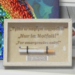 May include: A framed artwork with the text "For emergencies only!" in English, German, and Polish. The artwork features a cigarette, a match, and a matchbox. The frame is light-colored.