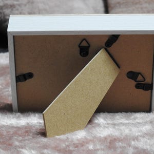 May include: Back view of a rectangular picture frame with a white painted wooden frame. The back is brown with black clips and a stand. The stand is made of cardboard. The frame is resting on a pink, fuzzy surface.