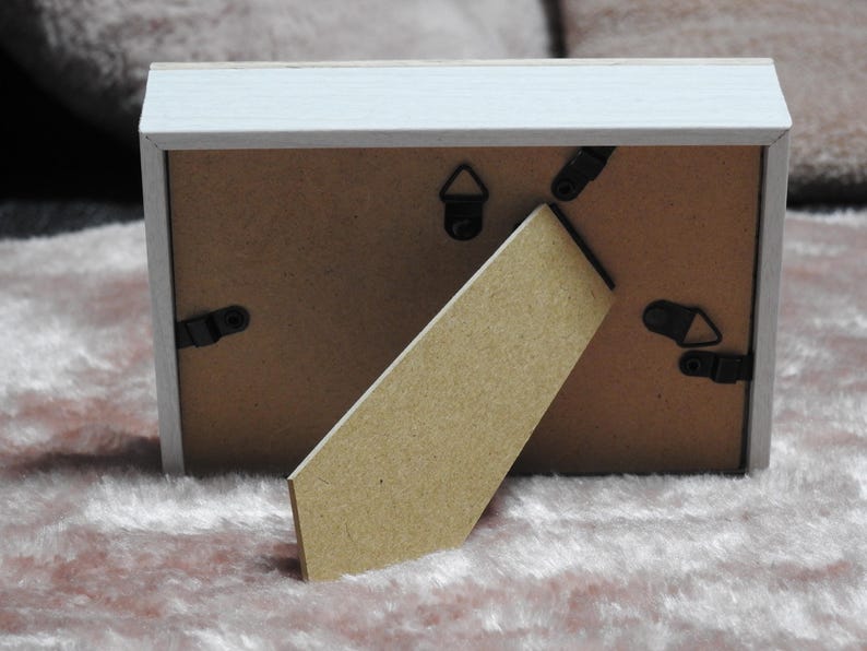 May include: Back view of a rectangular picture frame with a white painted wooden frame. The back is brown with black clips and a stand. The stand is made of cardboard. The frame is resting on a pink, fuzzy surface.