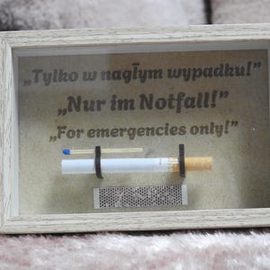 May include: A framed display with a cigarette, match, and matchbox. The text on the display reads "For emergencies only!" in English, German, and Polish. The frame is light-colored.