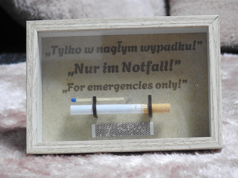May include: A framed display with a cigarette, match, and matchbox. The text on the display reads "For emergencies only!" in English, German, and Polish. The frame is light-colored.