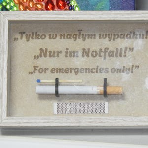 May include: A framed artwork with the text "For emergencies only!" in English, German, and Polish. The artwork includes a cigarette, a match, and a match strip, all held in place by black bands. The frame is white.