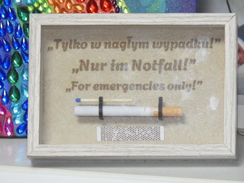 May include: A framed artwork with the text "For emergencies only!" in English, German, and Polish. The artwork includes a cigarette, a match, and a match strip, all held in place by black bands. The frame is white.