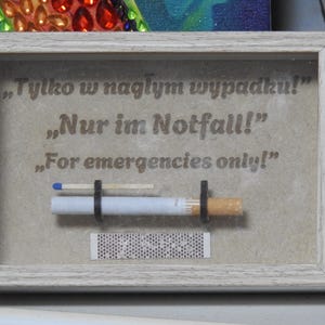 May include: A framed artwork with the text "For emergencies only!" in English, German, and Polish. The artwork features a cigarette, a match, and a matchbook, all held in place by black rubber bands. The frame is light gray.