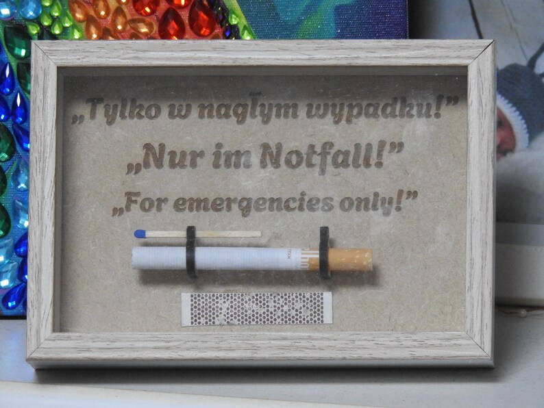 May include: A framed artwork with the text "For emergencies only!" in English, German, and Polish. The artwork features a cigarette, a match, and a matchbook, all held in place by black rubber bands. The frame is light gray.