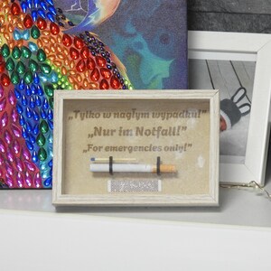 May include: A framed artwork with the text "For emergencies only!" in English, German, and Polish. The artwork features a cigarette, a match, and a matchbox. The frame is light-colored.