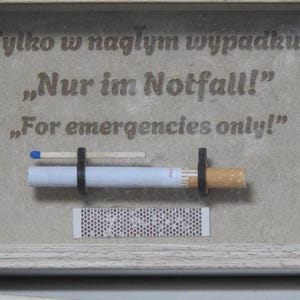 May include: A framed artwork with the text "For emergencies only!" in English, Polish, and German. The artwork features a cigarette, a match, and a matchbook. The frame is white.