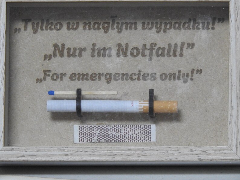 May include: A framed artwork with the text "For emergencies only!" in English, Polish, and German. The artwork features a cigarette, a match, and a matchbook. The frame is white.