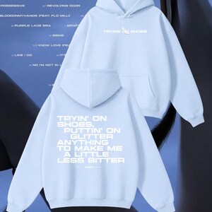 Könnte beinhalten: Ein hellblauer Kapuzenpullover mit einer Kängurutasche auf der Vorderseite. Auf der Brust steht der Text "TRYING ON SHOES" mit einem kleinen Symbol eines High-Heels. Auf der Rückseite befindet sich der weiße Text "TRYIN' ON SHOES, PUTTIN' ON GLITTER ANYTHING TO MAKE ME A LITTLE LESS BITTER."