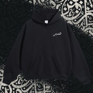 May include: A black hooded sweatshirt featuring a front kangaroo pocket and long sleeves. The chest area displays white Arabic calligraphy text that reads Sabr. The garment is constructed with a hood and ribbed cuffs at the wrists and hemline.