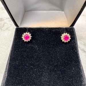 Puede incluir: Pendientes de botón con una piedra central rosa brillante rodeada por un halo de pequeñas piedras transparentes. Los pendientes están engarzados en metal dorado y presentados en un joyero de terciopelo negro.