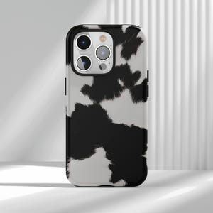 Könnte beinhalten: Eine Handyhülle mit Kuhfellmuster in Schwarz und Weiß. Die Hülle hat einen schwarzen Rand und eine glänzende Oberfläche. Das Design zeigt unregelmäßige schwarze Flecken auf weißem Hintergrund, die ein Kuhfellmuster imitieren. Die Handyhülle ist für ein Smartphone konzipiert.