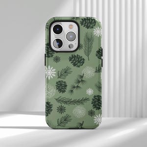 May include: A green phone case featuring a repeating pattern of pine cones, snowflakes, and pine branches. The design includes white and dark green elements, creating a winter or holiday theme. The case has a camera cutout and black side accents.