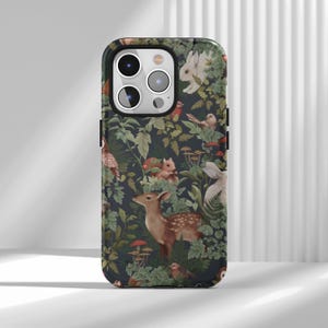 Puede incluir: Funda de teléfono con un diseño de bosque oscuro. El diseño presenta un ciervo, conejos, pájaros, ardillas y setas sobre un fondo de follaje verde. La funda tiene un borde negro y recortes para la cámara.