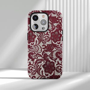 May include: A burgundy phone case featuring a detailed lace pattern. The design includes floral and swirling elements, creating a textured appearance. The case has a protective bumper and cutouts for the camera and buttons.