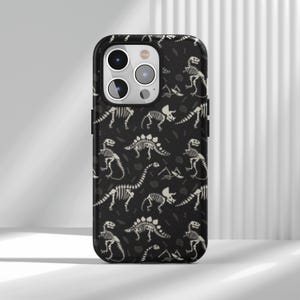 May include: Black phone case featuring a repeating pattern of white dinosaur skeletons. The design includes various dinosaur species and bone fragments. The case has a glossy finish and a black border.