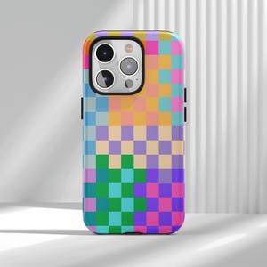 Puede incluir: Funda de teléfono colorida con un patrón de tablero de ajedrez en tonos rosa, morado, verde, azul y naranja. La funda tiene un acabado brillante y un borde negro alrededor de la cámara y los botones laterales.