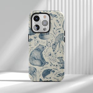 Op de afbeelding: Een telefoonhoesje met een crèmekleurige achtergrond en een herhalend patroon van blauwe bosdieren en planten. Het ontwerp omvat een beer, hert, vos, vogels, vlinders en een egel. De hoes heeft een zwarte rand en camera-uitsparingen.