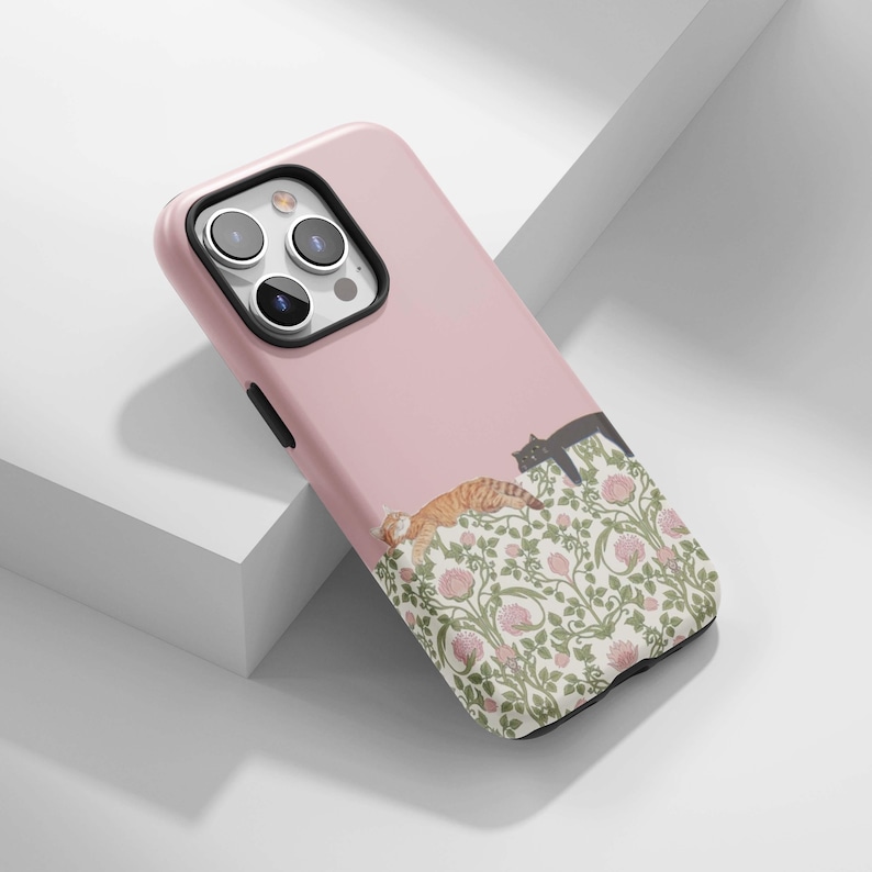 May include: A pink phone case featuring a floral design with two cats. One cat is orange and white, the other is black. The floral pattern includes pink flowers and green leaves. The case is designed for a smartphone.