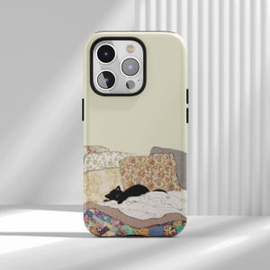 May include: A cream-colored smartphone case featuring a printed illustration of a small black cat lying on a bed with floral patterned pillows and a colorful patchwork quilt. The case has a cutout for a triple-lens camera system on the back.