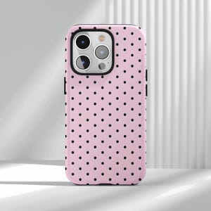 May include: Pink phone case with a black polka dot pattern. The case has a black border around the camera and the edges. The phone case is designed to fit a smartphone.