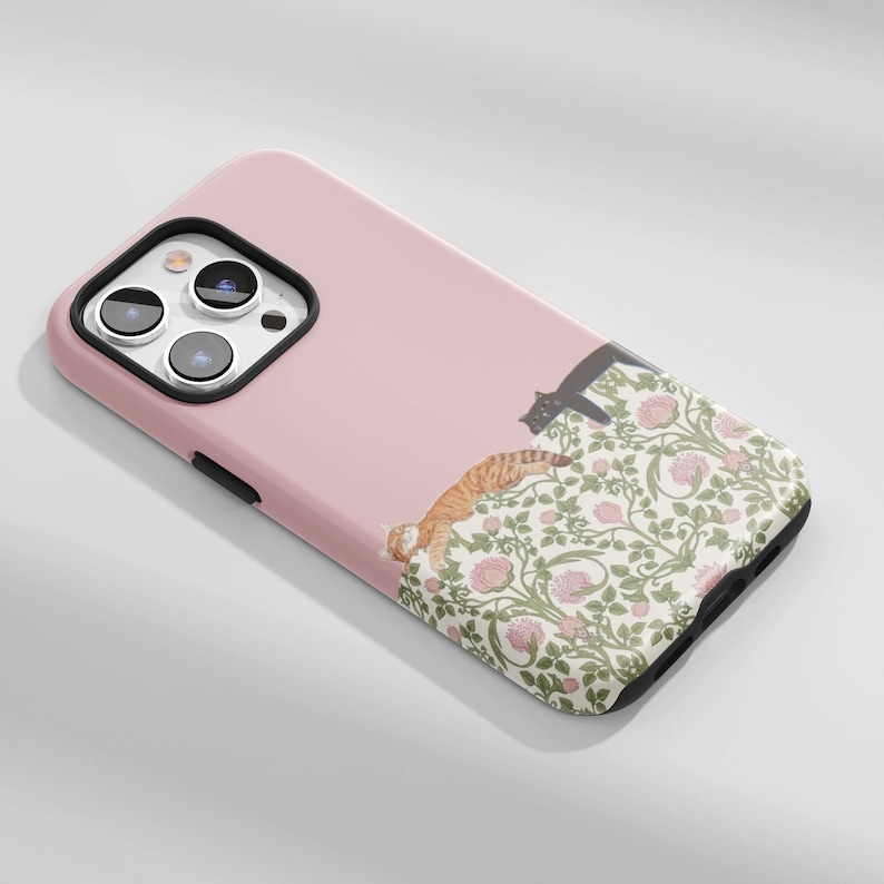 May include: A pink phone case featuring a floral pattern with pink roses and green leaves. Two cats are depicted on the case, one orange tabby and one black cat. The case has a camera cutout and black trim.