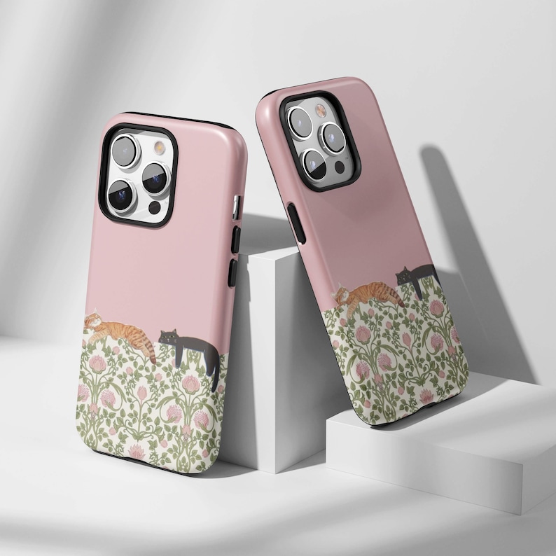 May include: Two pink phone cases with a floral design and sleeping cats. One case features an orange tabby cat and a black cat, while the other has a black cat and an orange tabby cat. The cases have black borders.