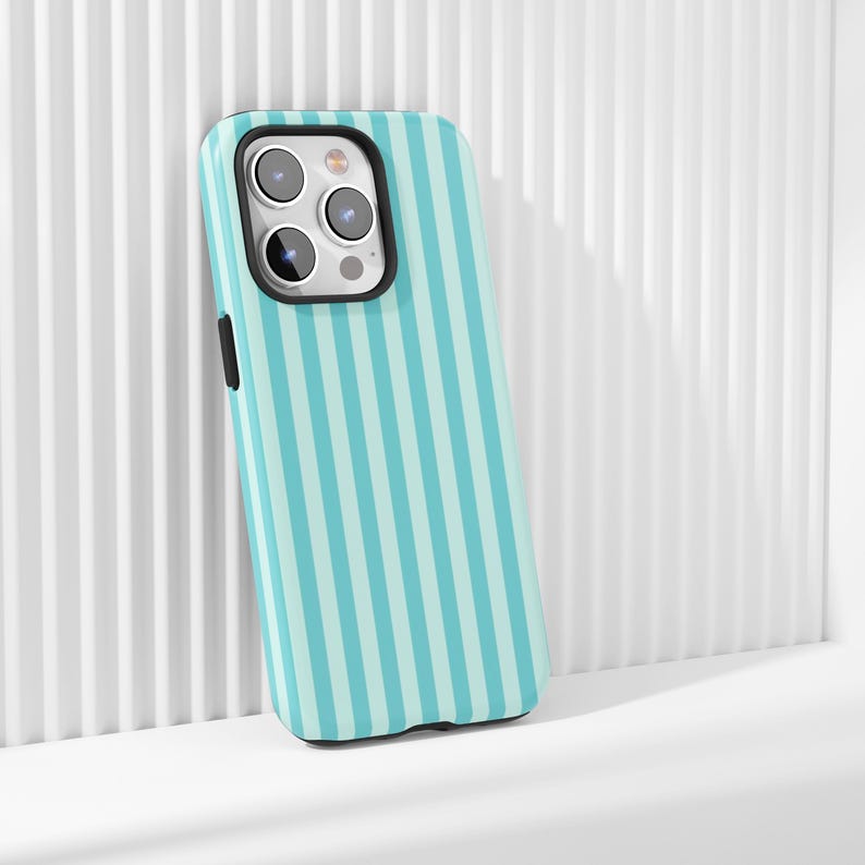 May include: A teal and light blue striped phone case with a black camera border. The case is leaning against a white, ridged surface. The phone case has a black side button.