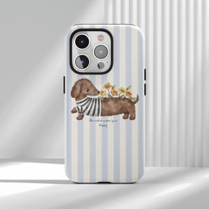 May include: A phone case with a light blue and white striped background. The case features a watercolor illustration of a brown dachshund wearing a striped shirt and carrying yellow flowers. The text "Do what makes you happy" is below the dog.