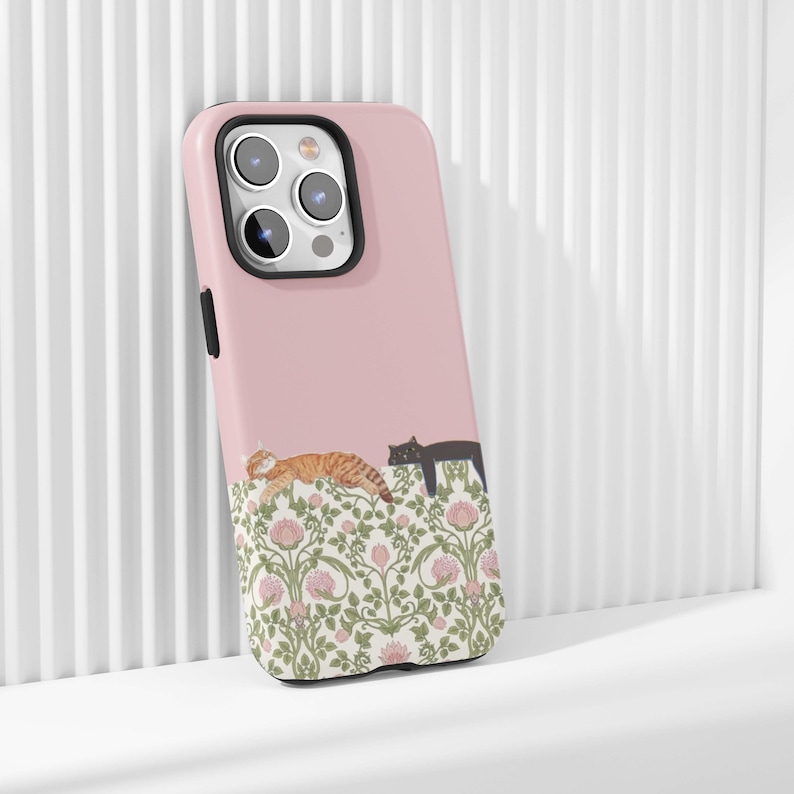 May include: A pink phone case featuring a design of two cats resting above a floral pattern. One cat is orange and the other is black. The floral pattern includes pink flowers and green leaves.