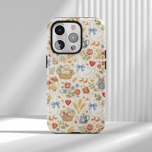 Pode incluir: Capa de telefone com um padrão caprichoso de gansos brancos com laços azuis, corações vermelhos, flores, cogumelos e uma chaleira. A capa tem um fundo creme e um acabamento brilhante, oferecendo uma estética vintage charmosa.