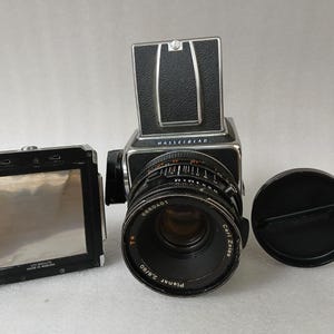 May include: A black Hasselblad medium format camera with a Carl Zeiss Planar 80mm lens and a lens cap. The camera has a silver top and a black leather grip. A separate black film back is also present.