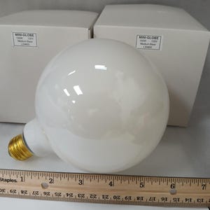 May include: A large, white, globe-shaped light bulb with a medium base, next to two white boxes. The bulb is approximately 6 inches in diameter. A wooden ruler is in the foreground.