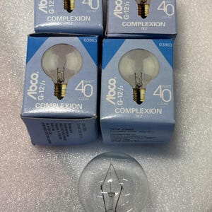 Könnte beinhalten: Eine Ansammlung von klaren Glühbirnen und Kartons. Die blauen Kartons tragen die Aufschrift "Abco G-12 1/2" und "40 Watt Clear". Die Glühbirnen haben eine runde Form mit einem Messingsockel. Der Text "COMPLEXION" ist ebenfalls sichtbar.