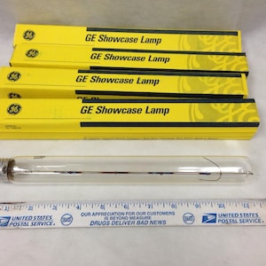 Op de afbeelding: Vier gele GE Showcase Lamp dozen met zwarte tekst en een heldere glazen lamp. Een liniaal met tekst van de United States Postal Service is ook zichtbaar. De lamp is ongeveer 30 cm lang.