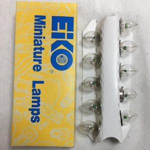 Könnte beinhalten: Eine gelbe Schachtel mit EIKO Miniaturlampen neben einer weißen Karte mit mehreren kleinen, klaren Glühbirnen. Die Schachtel hat blauen Text und ein Logo. Die Glühbirnen haben einen kleinen grünen Punkt im Inneren.