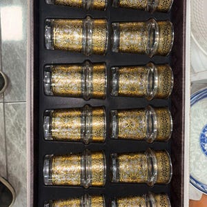 Puede incluir: Un juego de doce posavasos de vidrio transparente con intrincados diseños de filigrana dorada, dispuestos en una caja negra. Los posavasos tienen forma cilíndrica.