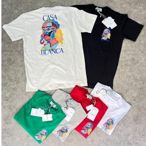 May include: A collection of Casa Blanca t-shirts in various colors, including white, black, green, gray, and red. Each shirt features a colorful graphic of a stylized face with the brand name. The shirts are short-sleeved and have tags attached.