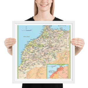 Op de afbeelding: Een ingelijste kaart van Marokko, met gedetailleerde geografische informatie. De kaart toont de Atlantische Oceaan en de Middellandse Zee, met een kleinere inzetkaart. De lijst is wit en de kaart heeft een groene rand.